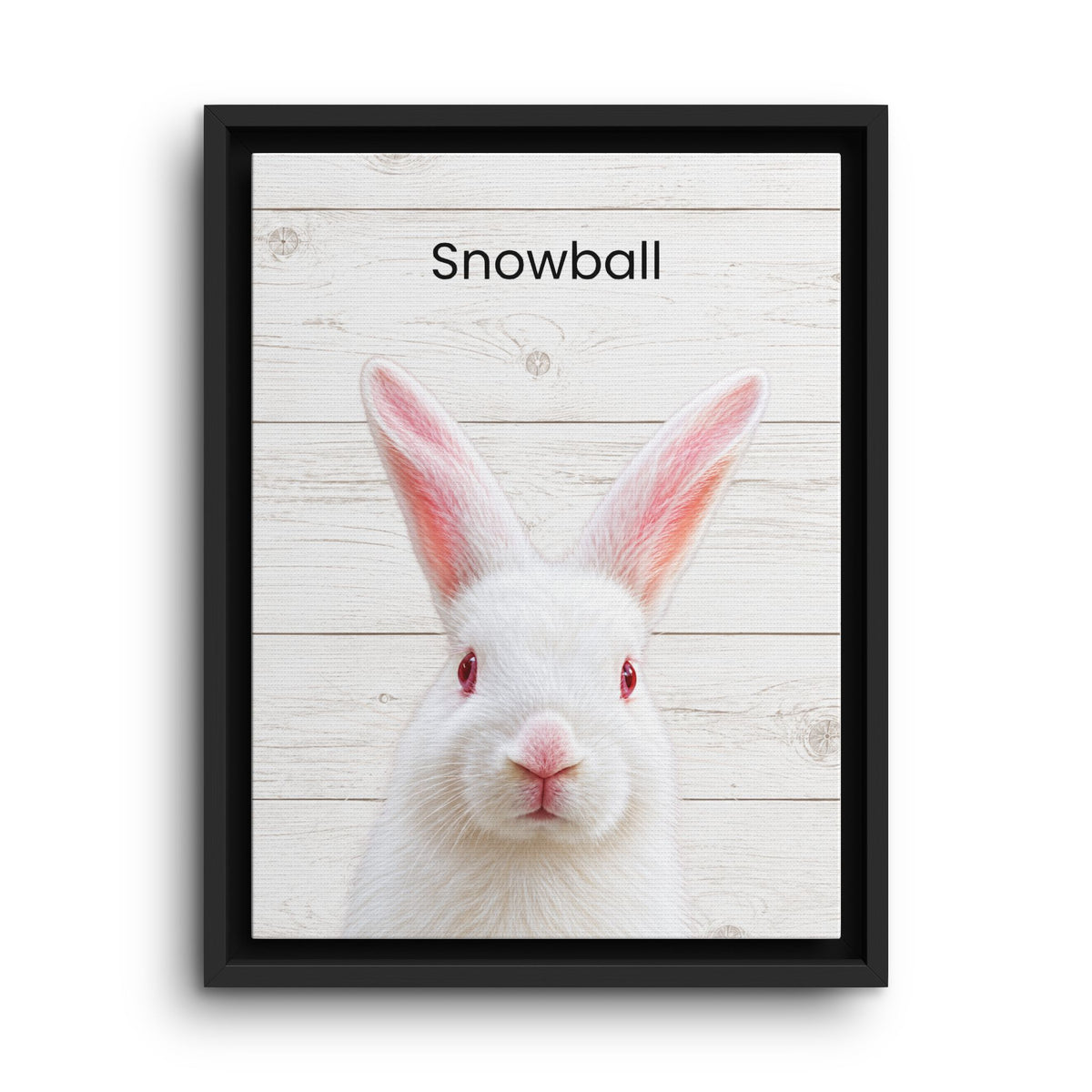 A framed art print featuring a white rabbit with red eyes and pink inner ears on a light wood plank background, with the word 'Snowball' printed at the top. The print is housed in a simple black frame and shown against a clean white background.