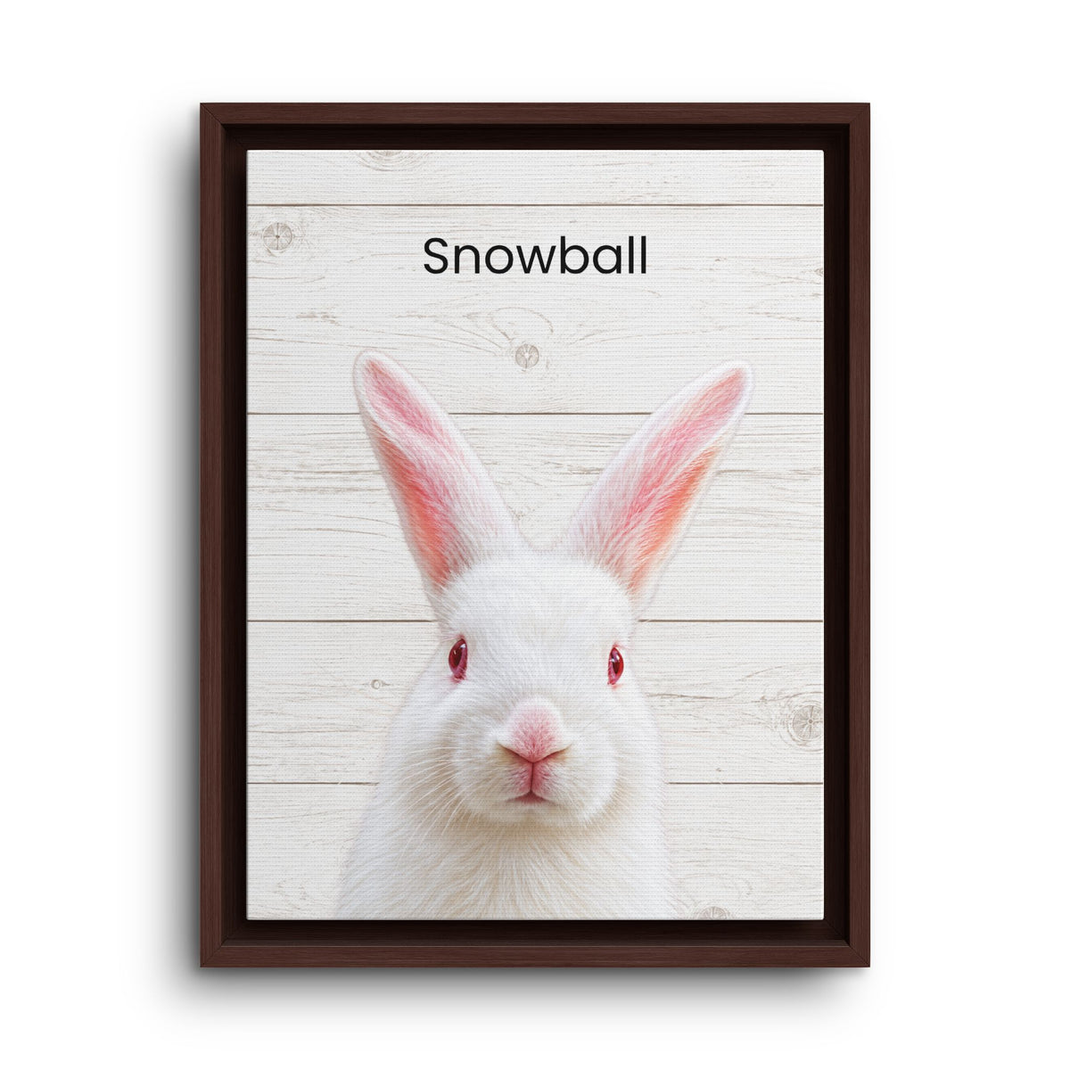 A framed art print featuring a white rabbit with red eyes and pink inner ears on a white wood plank background, with the word 'Snowball' printed at the top. The print is housed in a dark brown frame and shown against a plain white background.