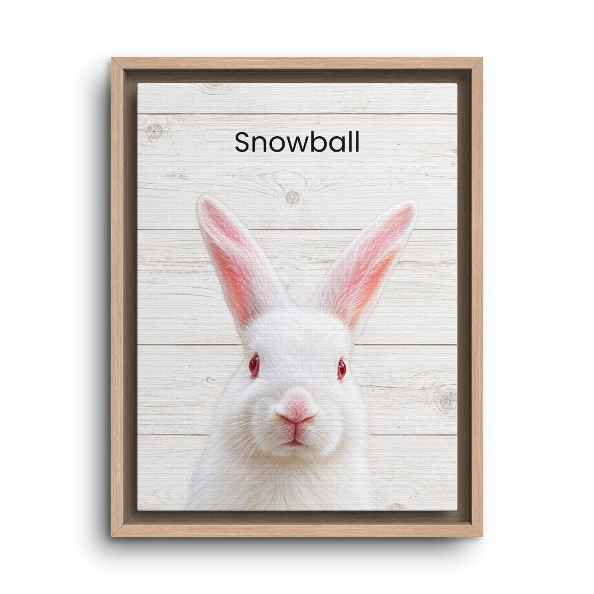 Framed wall art featuring a white rabbit centered on a whitewashed wood plank background, with the word 'Snowball' printed at the top. The piece is shown in a light wood frame against a plain white background.