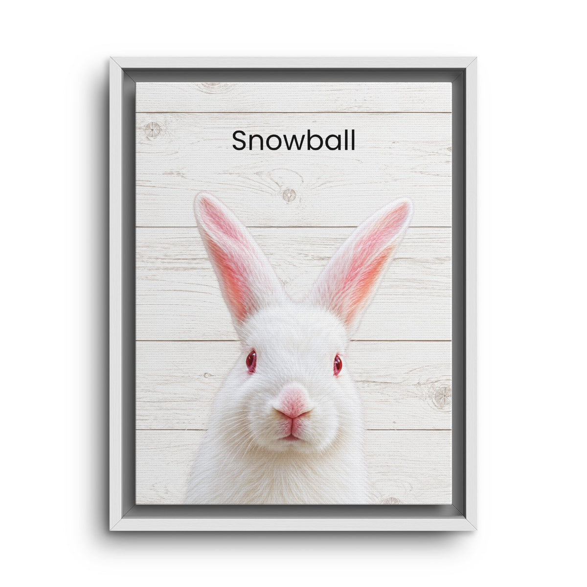 A framed custom pet portrait print featuring a white rabbit with red eyes and the name 'Snowball' printed at the top, set against a light wood plank background.