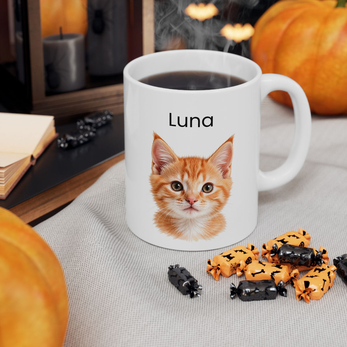 A white ceramic mug filled with black coffee sits on a light gray fabric surface, decorated with a printed orange tabby kitten face and the name 'Luna' above it. Pumpkins, wrapped candy, candles, and a book are visible in a cozy styled setting.