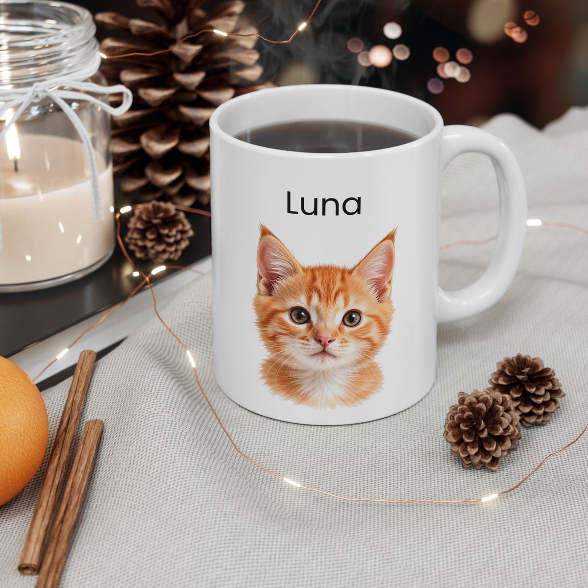 A white ceramic mug with a printed orange tabby kitten face and the name 'Luna' above it, filled with a dark beverage and styled on a light fabric surface with pinecones, fairy lights, cinnamon sticks, an orange, and a candle.