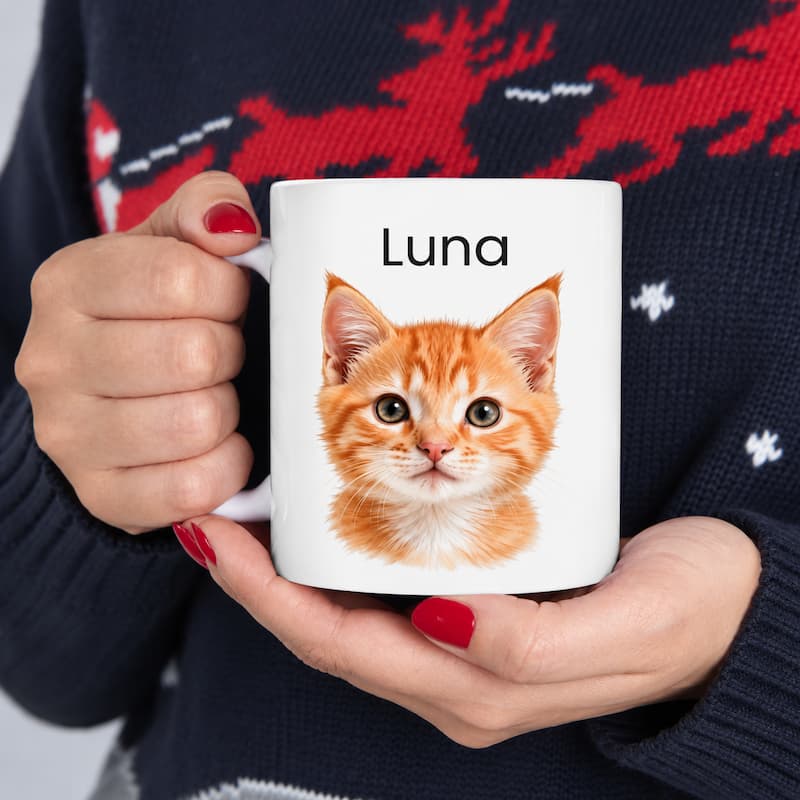 A person holding a white ceramic mug with a printed illustration of an orange tabby kitten and the name 'Luna' in black text. The person is wearing a dark navy knit sweater with red and white patterns.