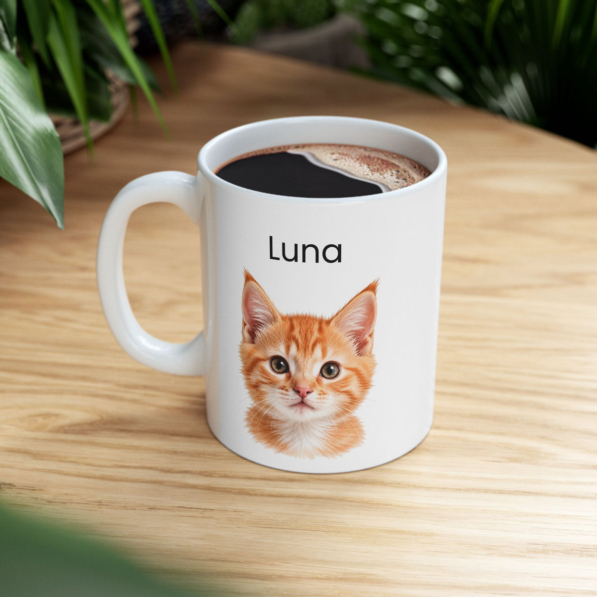 A white ceramic mug filled with black coffee sits on a light wood table. The mug is printed with the name 'Luna' above a detailed illustration of an orange tabby kitten.