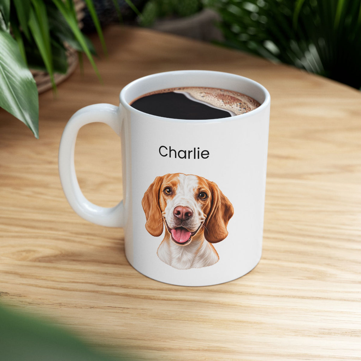 A white ceramic coffee mug with a printed portrait of a brown and white beagle dog and the name 'Charlie' above it, filled with black coffee and set on a light wood table with green plants in the background.