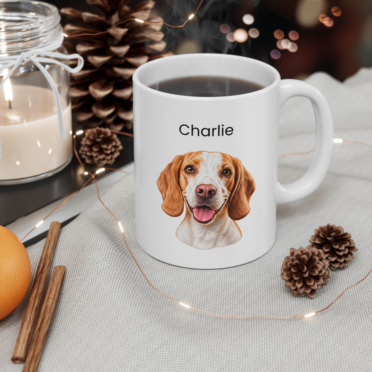 A white ceramic mug with a printed portrait of a brown and white beagle dog and the name 'Charlie' above it, shown on a light fabric surface with pinecones, a candle jar, cinnamon sticks, and string lights around it.